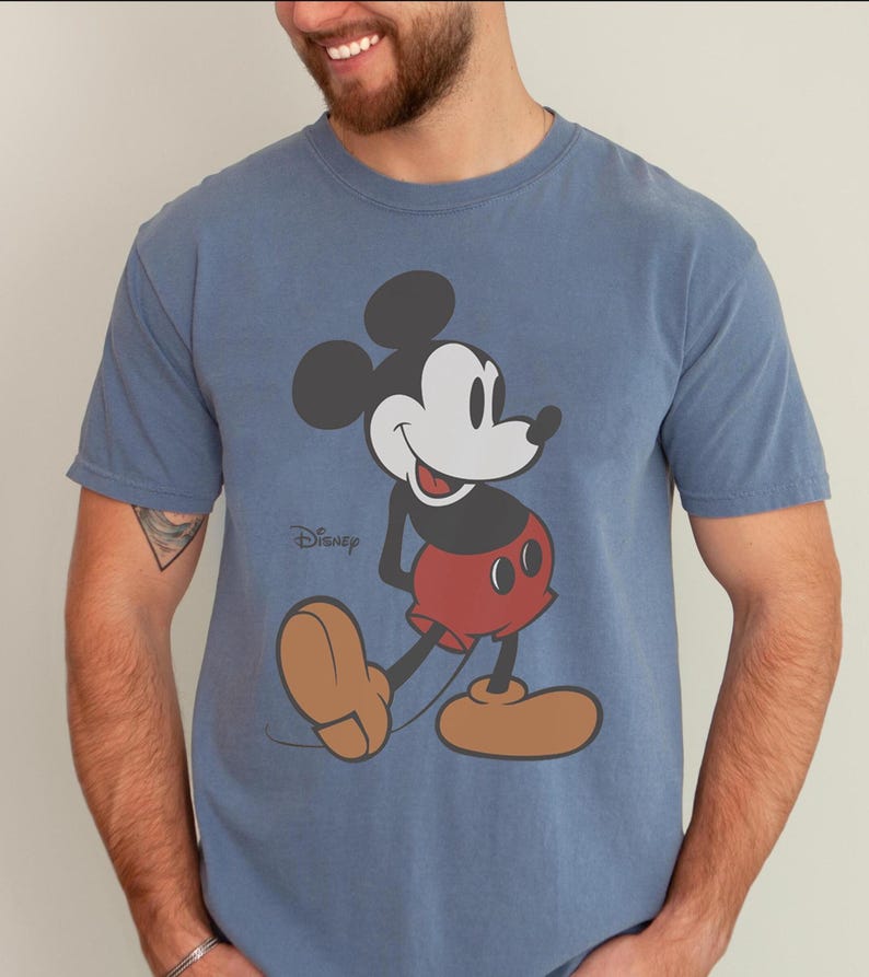May include: Blue t-shirt featuring a classic Mickey Mouse graphic. The cartoon character is depicted in black, white, red, and tan, with the Disney logo printed below. The shirt has short sleeves and a crew neck.