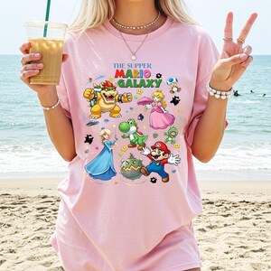 Puede incluir: Camiseta rosa claro con un gr&aacute;fico colorido de personajes de Super Mario Galaxy. El texto "THE SUPPER MARIO GALAXY" est&aacute; impreso encima de los personajes. La camiseta est&aacute; hecha de un material suave.