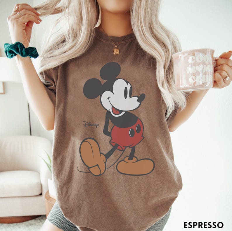 May include: Espresso-colored t-shirt featuring a classic Mickey Mouse graphic. The cartoon character is depicted in black, white, and red, with a smiling expression. The Disney logo is visible. The shirt is a casual wear item.