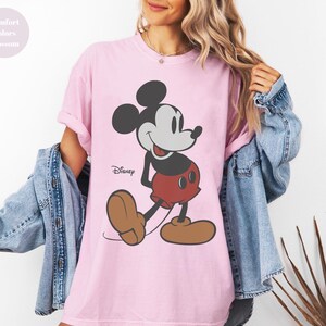 May include: A light pink t-shirt featuring a classic Mickey Mouse graphic. The cartoon character is depicted in black, gray, red, and brown. The Disney logo is printed below the character. The shirt is part of the Comfort Colors Blossom collection.