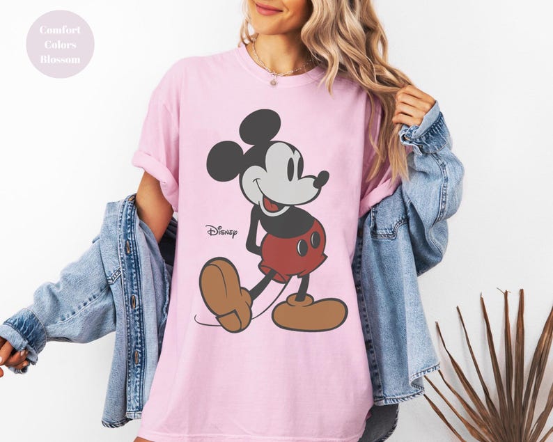 May include: A light pink t-shirt featuring a classic Mickey Mouse graphic. The cartoon character is depicted in black, gray, red, and brown. The Disney logo is printed below the character. The shirt is part of the Comfort Colors Blossom collection.