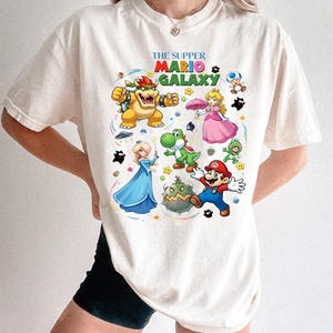 Puede incluir: Camiseta de color crema con un gr&aacute;fico colorido de personajes del videojuego Super Mario Galaxy. El dise&ntilde;o incluye a Mario, Bowser, la Princesa Peach, Yoshi y otros personajes, con el texto "THE SUPPER MARIO GALAXY".