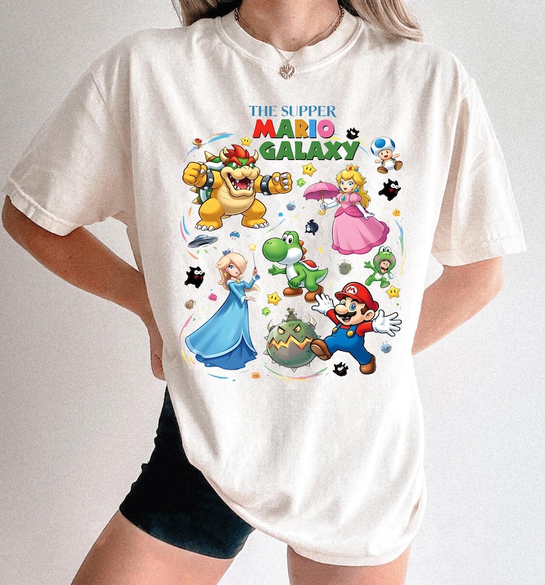 Puede incluir: Camiseta de color crema con un gr&aacute;fico colorido de personajes del videojuego Super Mario Galaxy. El dise&ntilde;o incluye a Mario, Bowser, la Princesa Peach, Yoshi y otros personajes, con el texto "THE SUPPER MARIO GALAXY".