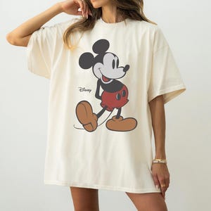 May include: Cream-colored oversized t-shirt featuring a classic Mickey Mouse graphic. The cartoon character is depicted in black, red, and brown, with the Disney logo printed below. The shirt has short sleeves and a relaxed fit, suitable for casual wear.