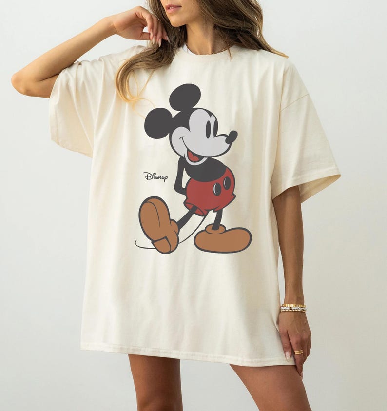 May include: Cream-colored oversized t-shirt featuring a classic Mickey Mouse graphic. The cartoon character is depicted in black, red, and brown, with the Disney logo printed below. The shirt has short sleeves and a relaxed fit, suitable for casual wear.