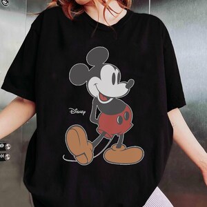May include: Black t-shirt featuring a large graphic of Mickey Mouse in his classic pose. The Disney logo is visible. Mickey Mouse is depicted with a white face, black ears, red shorts, and brown shoes. The shirt has short sleeves.