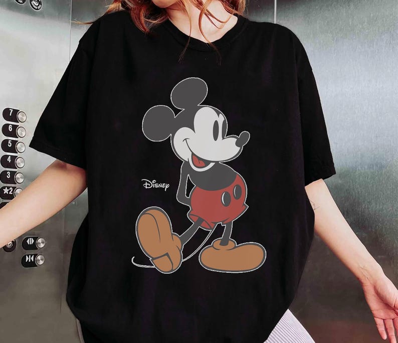 May include: Black t-shirt featuring a large graphic of Mickey Mouse in his classic pose. The Disney logo is visible. Mickey Mouse is depicted with a white face, black ears, red shorts, and brown shoes. The shirt has short sleeves.