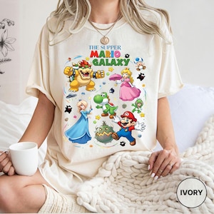 Puede incluir: Camiseta de color marfil con un gr&aacute;fico colorido de personajes del videojuego Super Mario Galaxy. El dise&ntilde;o incluye a Mario, la Princesa Peach, Bowser, Yoshi y otros personajes. El texto "THE SUPPER MARIO GALAXY" est&aacute; impreso en la parte superior.