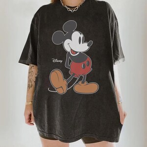 May include: Dark gray oversized t-shirt featuring a classic Mickey Mouse graphic. The cartoon character is depicted in black, red, and tan colors, with the Disney logo printed on the shirt. The shirt has short sleeves and a relaxed fit.