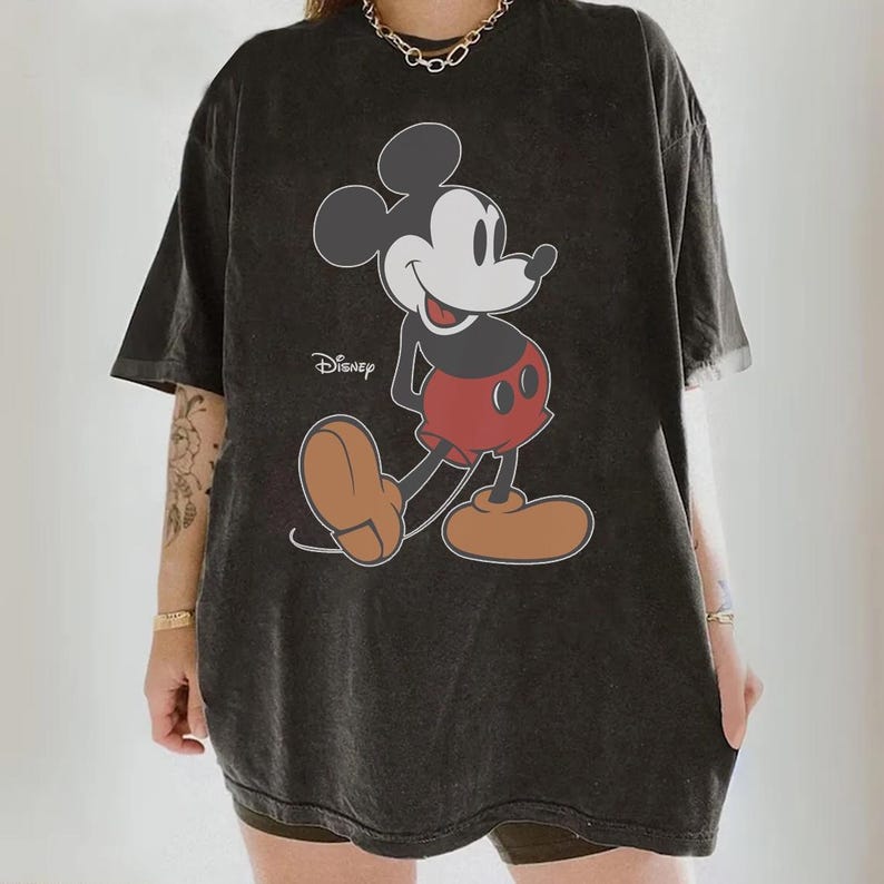 May include: Dark gray oversized t-shirt featuring a classic Mickey Mouse graphic. The cartoon character is depicted in black, red, and tan colors, with the Disney logo printed on the shirt. The shirt has short sleeves and a relaxed fit.
