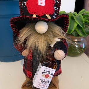 May include: A handmade gnome figurine wearing a red and black plaid jacket and hat with the word "BEAM" on it. The gnome has a long, fluffy beard and is holding a miniature bottle of Jim Beam bourbon.