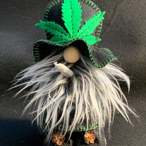 May include: A novelty gnome figure with a long, grey and white beard, sporting a black hat featuring a large green cannabis leaf. The gnome holds a small, rolled item, and its black boots are decorated with small, brown accents.
