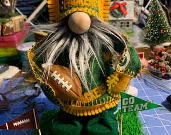 May include: Green Bay Packers gnome with a white beard, wearing a green and yellow jersey with the team logo and holding a football. The gnome is standing on a green grass surface with a green and white flag that says "GO TEAM".