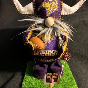 May include: A purple and gold gnome wearing a Minnesota Vikings hat and jersey, holding a football. The gnome is standing on a green grass mat with a sign that says "Touch Down!" and a number one sign.