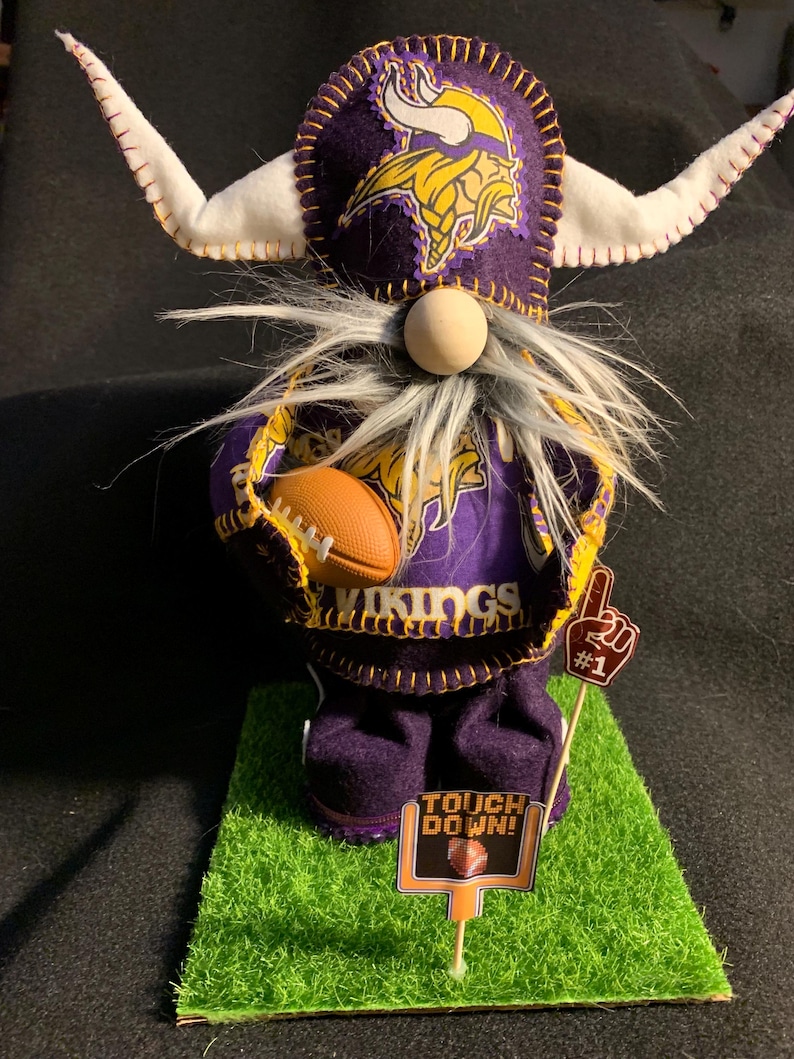 May include: A purple and gold gnome wearing a Minnesota Vikings hat and jersey, holding a football. The gnome is standing on a green grass mat with a sign that says "Touch Down!" and a number one sign.
