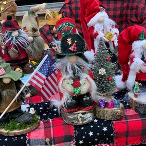 May include: A collection of handmade gnomes, some dressed in red and white, others in green and brown, with a small American flag and a miniature Christmas tree. The gnomes are displayed on wooden rounds with moss and are arranged on a red and black plaid fabric.