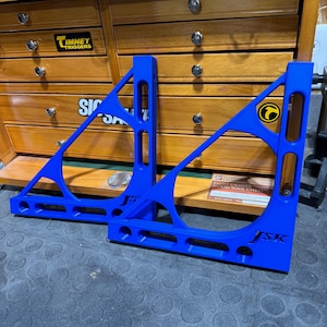 Puede incluir: Dos escuadras de esquina de metal azul con el texto "JSK" impreso en ellas. Las escuadras están sobre una superficie negra.