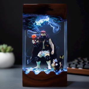 Puede incluir: Una pieza de arte en resina que presenta una figura musculosa con una antorcha en llamas, un gato y un fondo azul ahumado. La figura lleva una capa negra, rodilleras y ropa interior con estampado de corazones. La pieza está encerrada en un marco de madera rectangular.