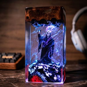K&ouml;nnte beinhalten: Ein rechteckiges Kunstwerk aus Kunstharz mit einer schwarzen Figur des Ghostface-Killers aus den Scream-Filmen, beleuchtet von blauen und violetten Lichtern. Die Oberseite ist aus dunklem Holz, die Basis hat rote und wei&szlig;e Akzente.