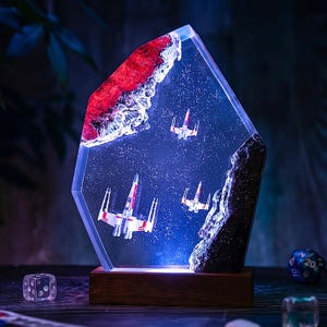 以下が含まれることがあります： 青く照らされた宇宙空間に3機のXウイング・スターファイターのミニチュアが浮かぶ、樹脂製の装飾ランプ。ギザギザとした結晶のような形状で、赤と黒の質感のある縁取りが施され、長方形の木製ベースに取り付けられている。