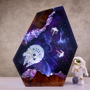 Op de afbeelding: Een zeshoekig harskunstwerk met een ruimtescène met een Millennium Falcon en TIE-jagers. Het kunstwerk heeft een bruine basis en een paars-blauw galaxy-ontwerp. Een klein astronautenfiguur staat ernaast.