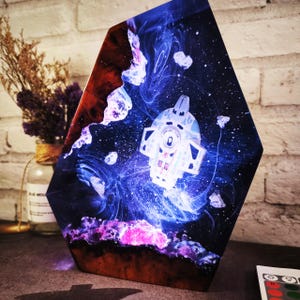 Pode incluir: Uma lâmpada hexagonal de resina com um design temático espacial. A lâmpada apresenta um elemento branco brilhante em forma de nave espacial no centro, rodeado por padrões rodopiantes azuis e roxos e formas brancas flutuantes. A base é de cor castanho escuro e vermelho.