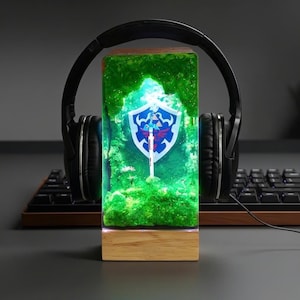 May include: A rectangular wooden-based lamp featuring a green, glowing forest scene with a shield and sword emblem. Black headphones rest on the lamp, which is placed in front of a keyboard. The lamp's design is inspired by a video game.