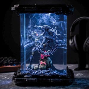 May include: A rectangular resin display case featuring a dark, biomechanical creature figure suspended within a clear block. The interior is filled with swirling white smoke effects, illuminated by a blue light, showcasing the detailed alien form.