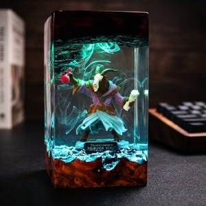 May include: A rectangular resin art piece featuring a goat-like figure in a purple robe, holding a red vial and a white object. The figure is encased in clear resin with blue and green swirling effects. The base is a dark wood.