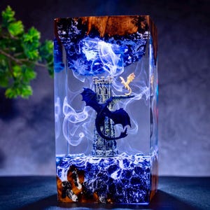 May include: A rectangular resin lamp featuring a dark blue dragon perched on a stone tower and a smaller light-colored dragon above it. The clear resin contains swirling white smoke effects, with natural wood grain at the top and bottom edges.