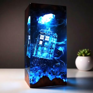 May include: A rectangular resin lamp featuring a blue police box design, with the text "POLICE BOX" visible. The lamp has a wooden base and is illuminated with blue light, creating a glowing effect. The overall theme is science fiction.