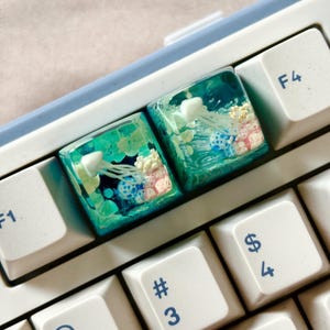 Puede incluir: Dos teclas de teclado personalizadas con un diseño de resina azul y verde que presenta medusas y corales. Las teclas están en un teclado blanco.
