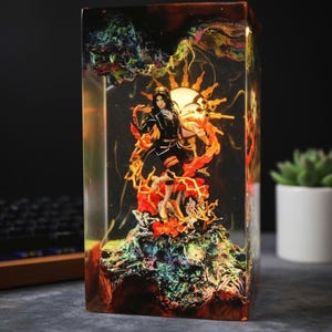 Peut inclure: Une sculpture rectangulaire en résine représentant une figurine féminine vêtue d'une tenue noire et orange, devant un motif en forme de soleil. La sculpture est enfermée dans un bloc de résine transparent avec des accents colorés et tourbillonnants et un fond sombre.