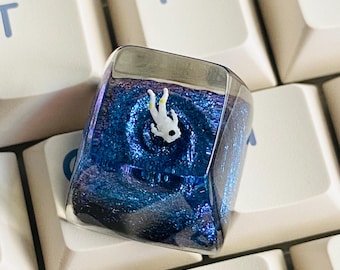 Peut inclure: Un keycap personnalisé avec un astronaute miniature flottant dans un tourbillon bleu et violet. Le keycap carré a une finition claire et brillante, sur fond de touches de clavier blanches.