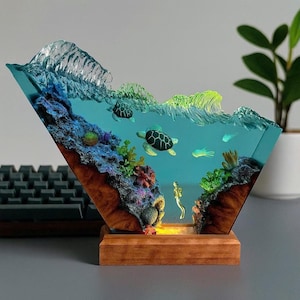 May include: A decorative resin sculpture depicting an underwater scene with sea turtles, coral, and a diver. The sculpture is encased in a clear, angled block, with a wooden base. The scene is illuminated from below, creating a warm glow.