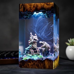 Puede incluir: Lámpara rectangular de resina con una escena prehistórica. Tiene una base y tapa de madera, con patrones arremolinados azules y blancos en el interior. La escena muestra figuras de dinosaurios y una estructura en miniatura.