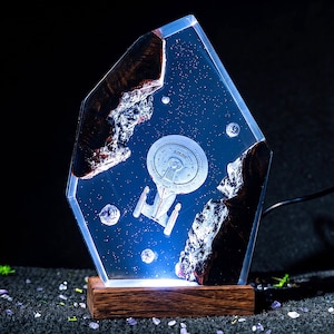 May include: A hexagonal resin lamp featuring a miniature Starship Enterprise model suspended within a clear blue space-themed interior. The piece is framed by two dark, textured wood segments and rests on a rectangular wooden base with an integrated light.