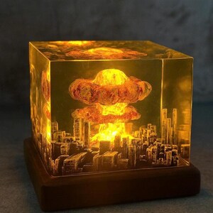 May include: A glowing, amber-colored cube lamp featuring a detailed cityscape silhouette at the base and a mushroom cloud design in the center. The lamp sits on a dark brown wooden base, creating a dramatic effect.