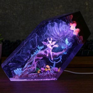 May include: A glowing resin sculpture featuring a fantasy creature perched on a branch. The artwork is encased in a clear, angled block, with a purple and blue color scheme. The scene includes a tree, flowers, and a starry background.
