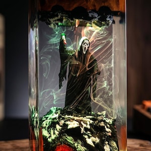 K&ouml;nnte beinhalten: Ein Kunstwerk aus Kunstharz mit einer Scream-Figur, die ein gr&uuml;nes Licht h&auml;lt. Die Figur ist in einem transparenten rechteckigen Block mit farbigen Wirbeln und einer felsigen Basis eingeschlossen. Die Oberseite hat dunkle, strukturierte Elemente.