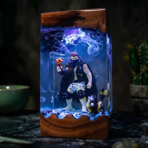 Op de afbeelding: Een rechthoekige houten lamp met een kern van heldere hars die blauw verlicht is. Binnenin staat een mannelijk actiefiguur met een zwarte cape en ondergoed met hartjespatroon naast een klein kattenfiguurtje in een wolk van rook.