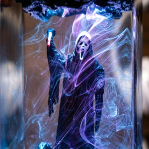 K&ouml;nnte beinhalten: Eine schwarze Ghostface-Figur aus dem Film Scream, in einem transparenten, rechteckigen Beh&auml;lter. Die Figur ist von wirbelnden blauen und violetten Lichtspuren umgeben, die einen unheimlichen, &auml;therischen Effekt erzeugen.