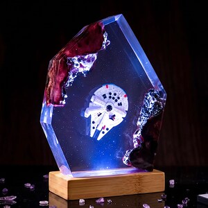 Op de afbeelding: Een sculptuur van heldere hars met een ingebed wit miniatuur ruimteschipmodel, omringd door een diepblauw sterreneffect. De hars is omlijst door donkere, getextureerde houten elementen en rust op een rechthoekige, lichtgekleurde houten voet.