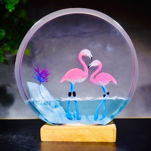Può includere: Un espositore circolare in resina trasparente con due statuette di fenicotteri rosa in piedi nell'acqua blu. A sinistra si trova una formazione rocciosa azzurra con una piccola pianta viola. L'oggetto è montato su una base in legno chiaro.