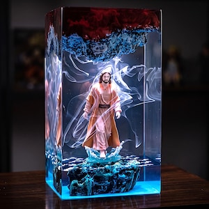 May include: A rectangular resin block containing a figurine of a person with long hair and a robe standing on a dark rock formation. The scene features swirling white smoke-like patterns, a blue water splash effect, and a dark red top section.