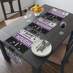 Puede incluir: Un camino de mesa con el texto "I AM ACE ENOUGH!" en blanco y morado sobre un fondo negro y gris. El camino de mesa está decorado con símbolos de naipes. Un plato blanco con tenedores y un cuenco de manzanas están sobre la mesa.