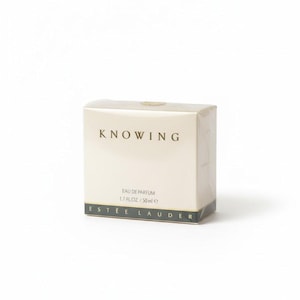 May include: A rectangular perfume box with the word "KNOWING" in a serif font. The box is off-white with gold and black accents. The text "EAU DE PARFUM 1.7 FL.OZ. / 50 ml e" is printed on the box.