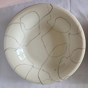 May include: A cream-colored ceramic bowl with a black abstract line design. The bowl has a rounded shape and a smooth surface. The black lines create an organic, free-flowing pattern across the interior.