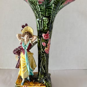 May include: A vintage ceramic vase featuring a woman in a yellow dress and a floral design. The vase is green with pink flowers and a flared top. The woman figure is wearing a hat and has her hands behind her head. The vase is likely around 25 cm tall.