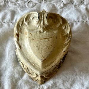 May include: Cream-colored, ornate wall sconce with a shield design. The sconce has a distressed finish, with visible wear and tear, and is made of a solid material. The design features decorative scrollwork and a small loop at the top for hanging.